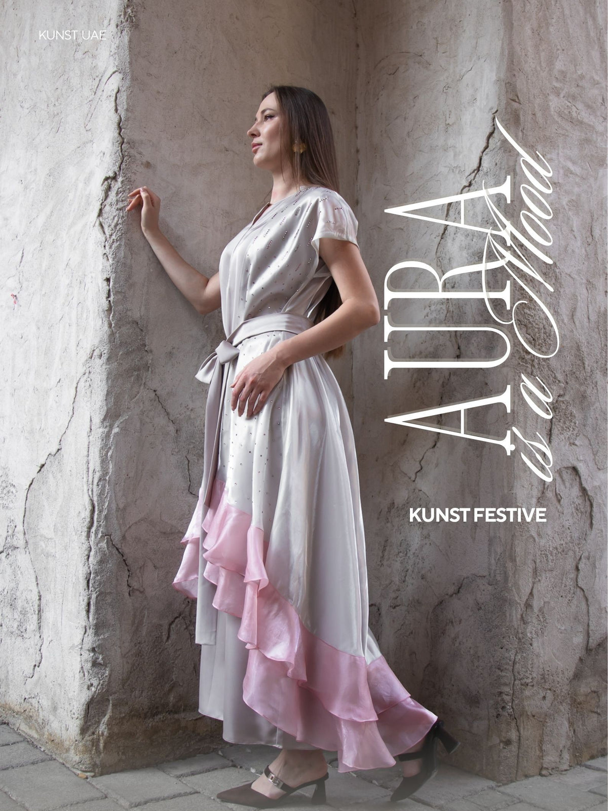 Woman in a grey and pink gown with a ruffled hem, standing against a stone wall.